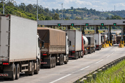 Fila de caminhões aguarda passagem pelas cabines de uma praça de pedágio durante o dia, com vegetação ao redor.