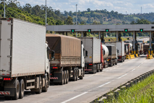 Fila de caminhões aguarda passagem pelas cabines de uma praça de pedágio durante o dia, com vegetação ao redor.