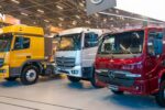 Três caminhões Mercedes-Benz, nas cores amarela, branca e vermelha, expostos lado a lado em um pavilhão da Fenatran