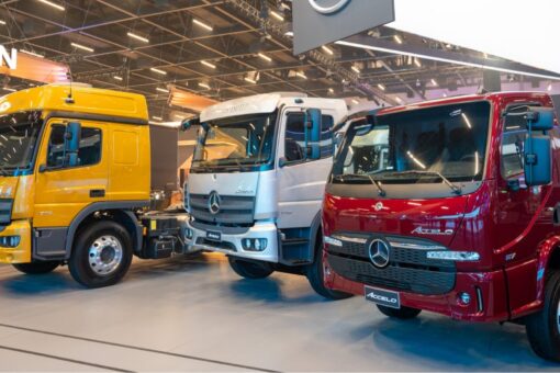 Três caminhões Mercedes-Benz, nas cores amarela, branca e vermelha, expostos lado a lado em um pavilhão da Fenatran